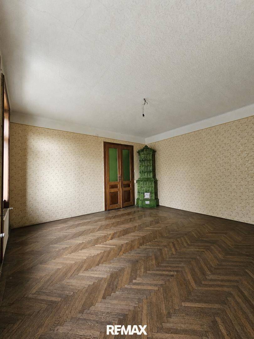 Geräumiges Zimmer mit Fischgrätparkett, gemusterter Tapete und einem traditionellen grünen Kachelofen.