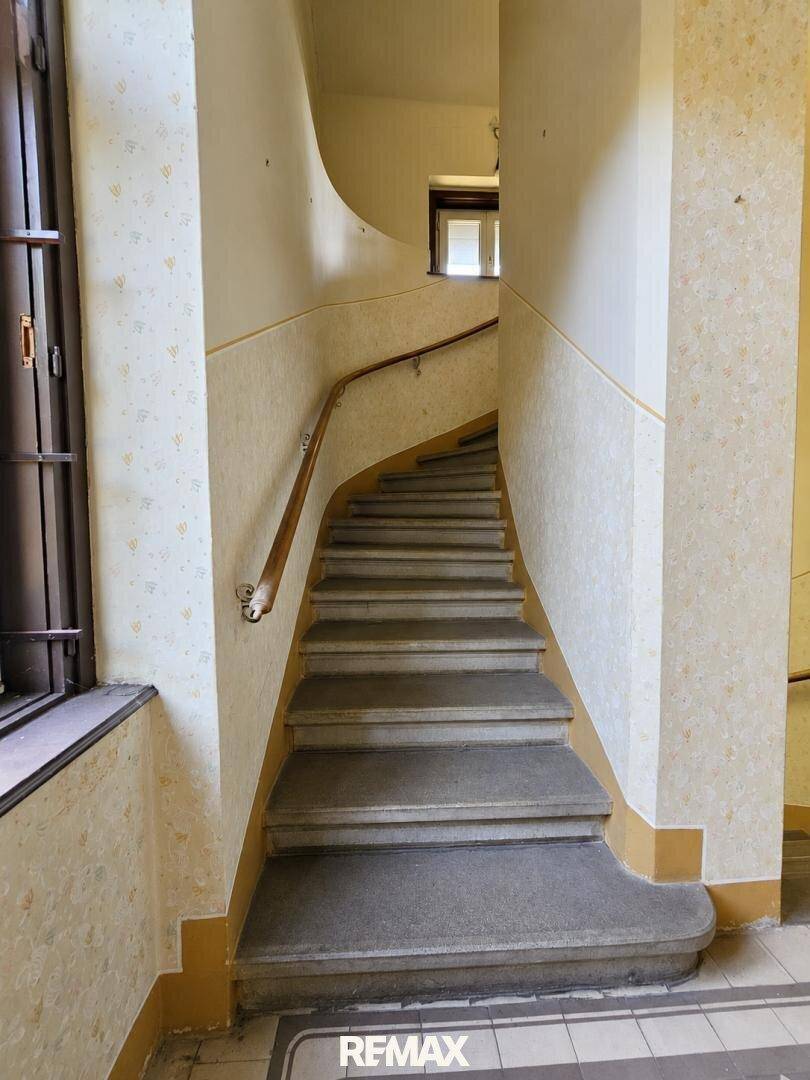 Altes Treppenhaus mit gemusterter Tapete, Holzgeländer und Stufen in originalem Zustand.
