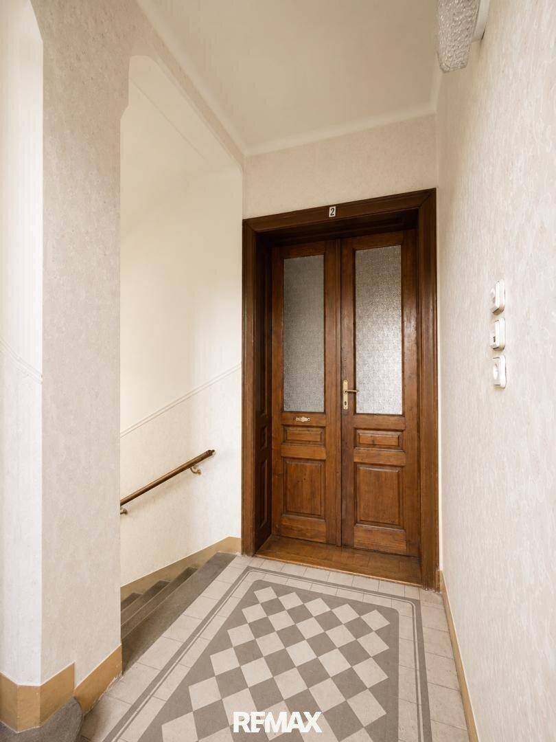 Eingangsbereich mit einer alten Holztür, gemustertem Fliesenboden und Tapeten im Treppenhaus.