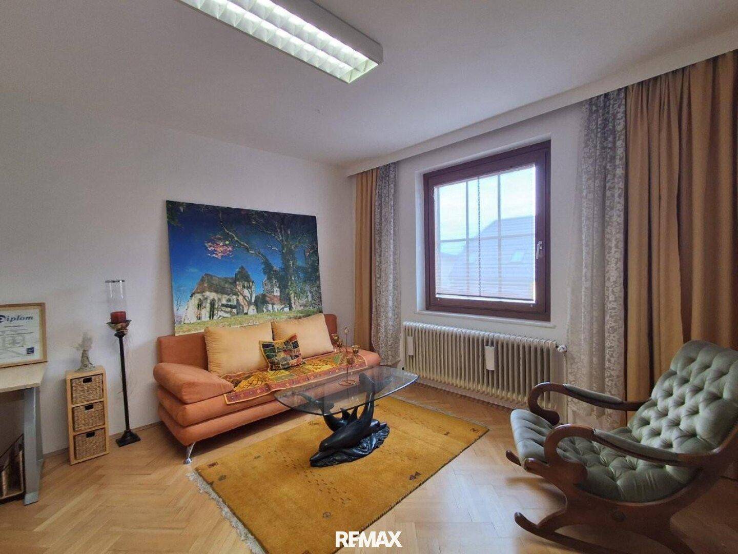 Helles Wohnzimmer mit Parkettboden, beiger Couch und großem Fenster mit Heizkörper.