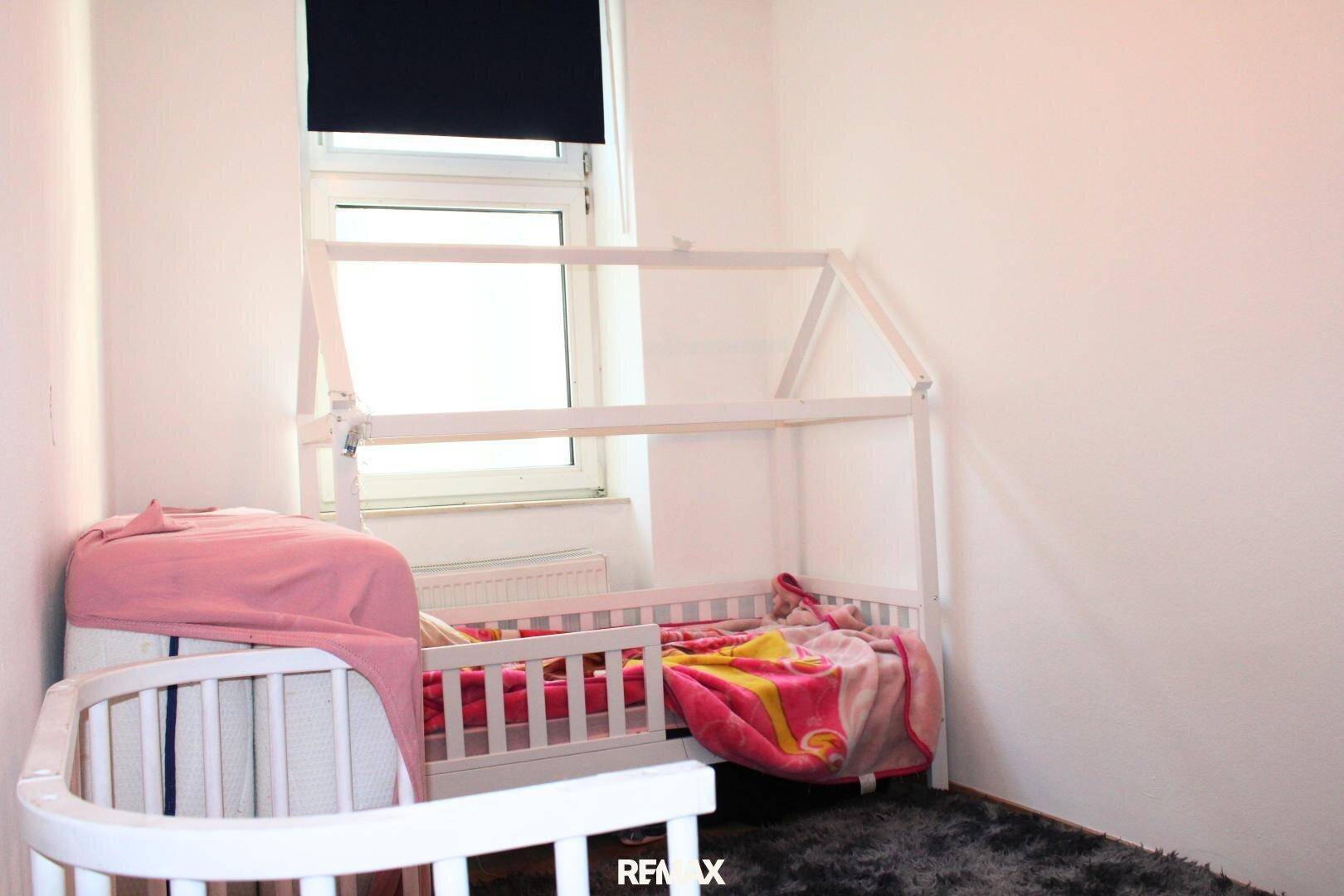 Kinderzimmer mit einem kleinen Hausbett, Babybett und einem Fenster.
