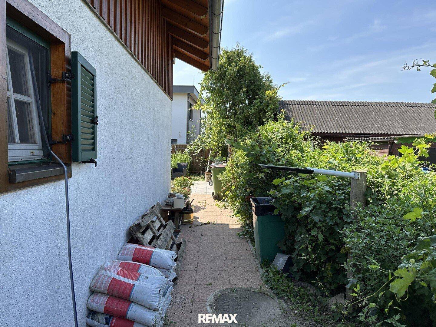Seitenansicht des Hauses mit schmalem Weg, Sträuchern und Mülltonnen.
