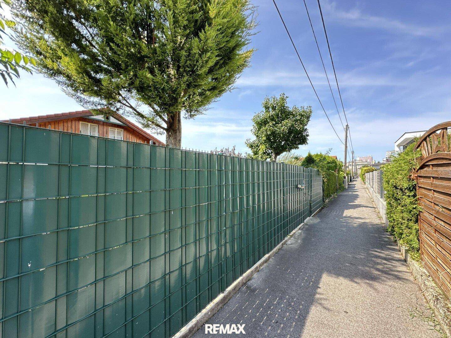 Schmaler Weg entlang eines grünen Zauns mit Bäumen und Häusern im Hintergrund.