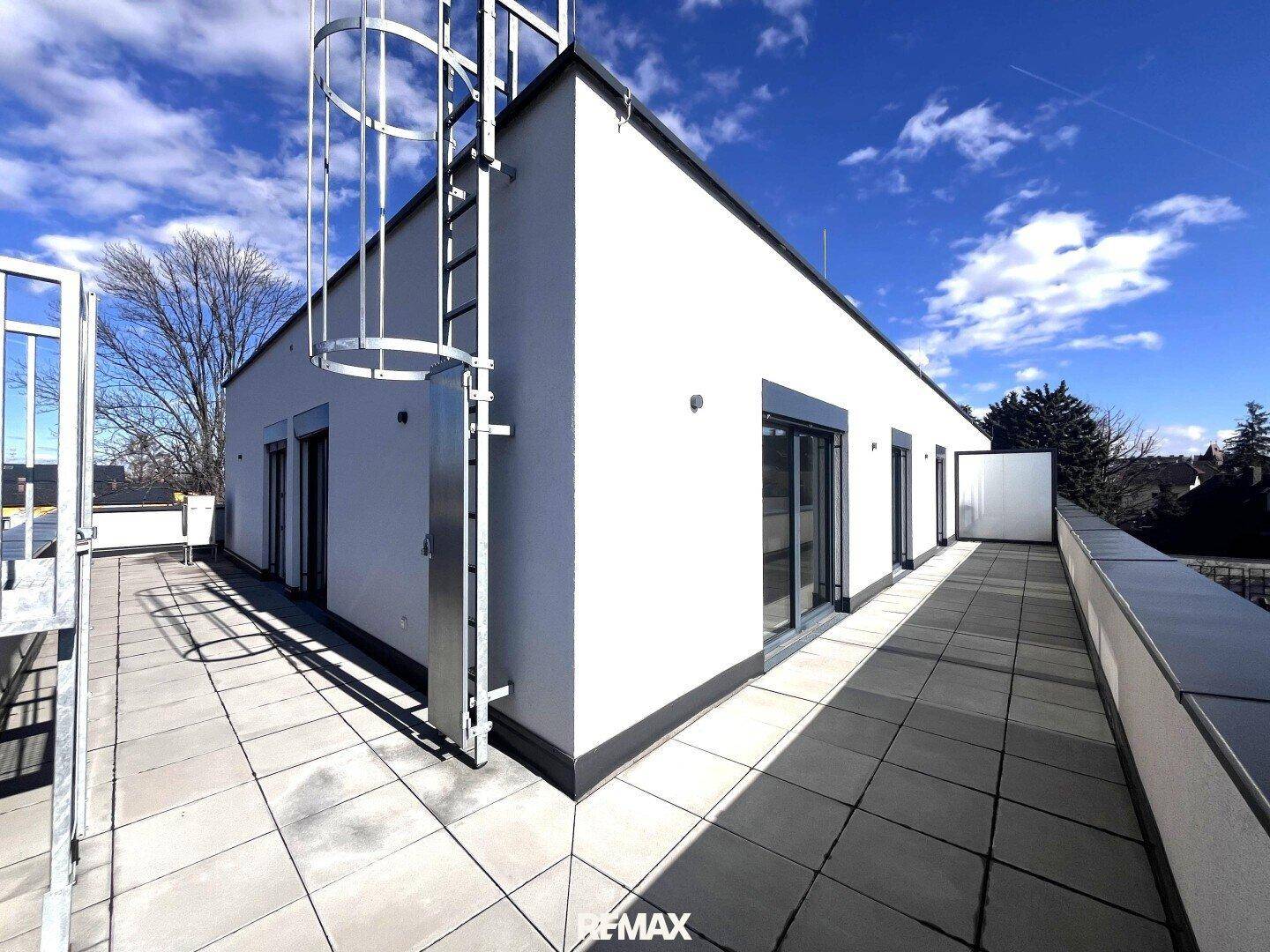 Große Dachterrasse mit Fliesenboden und Zugang zu den Wohnräumen unter blauem Himmel.