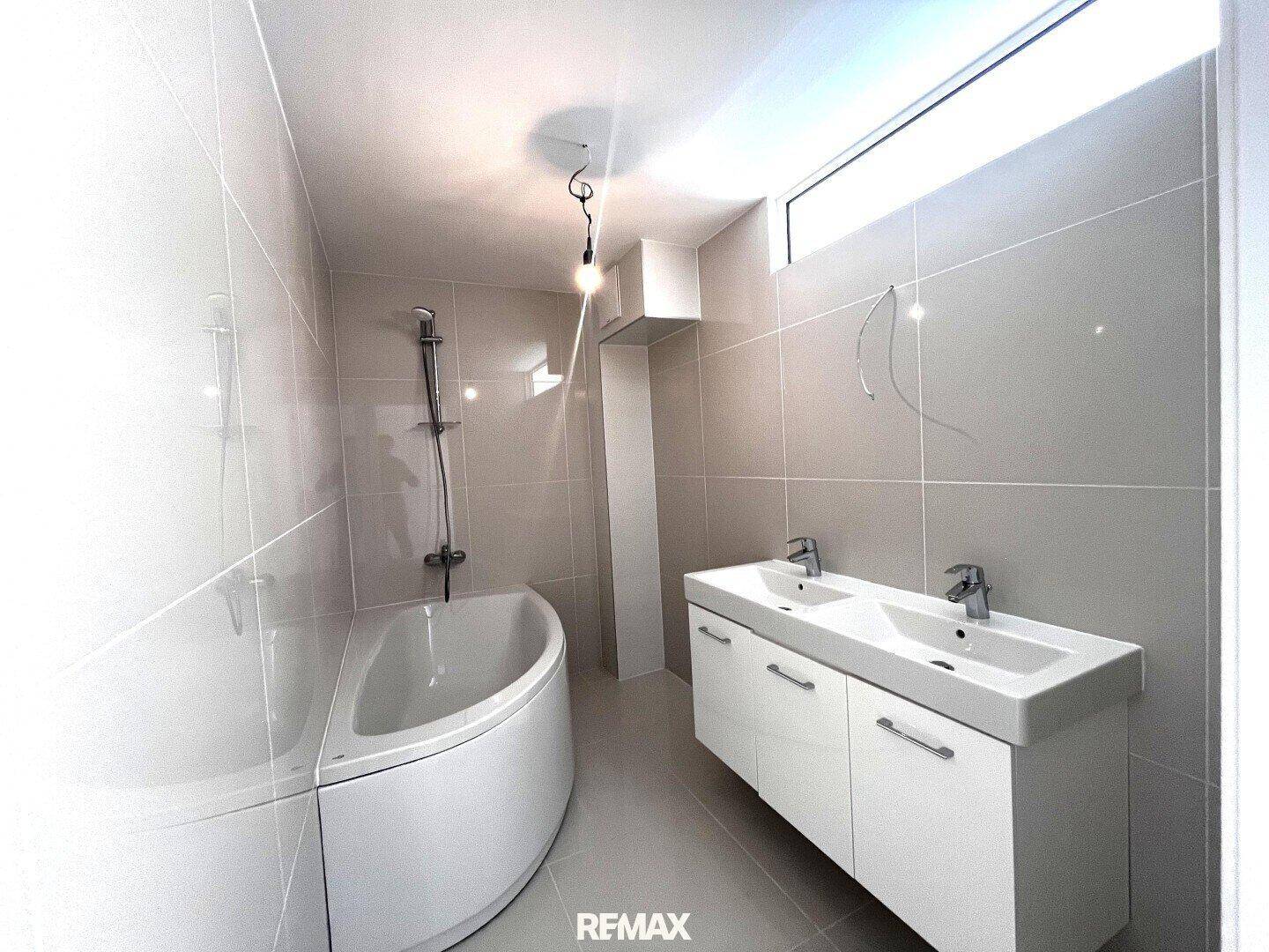 Modernes Badezimmer mit hellgrauen Fliesen, Eckbadewanne und Doppelwaschbecken.