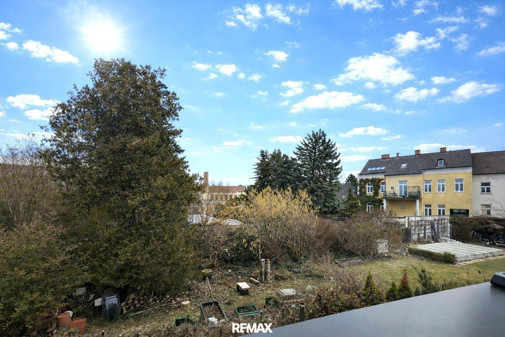 Ausblick auf einen grünen Gartenbereich mit Bäumen und angrenzenden Häusern unter blauem Himmel.