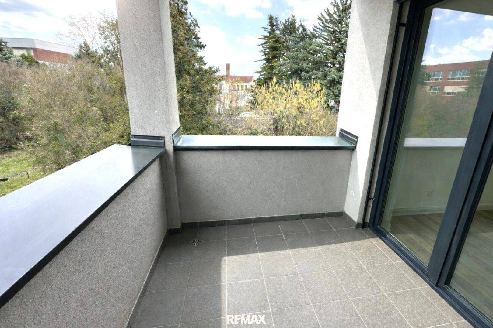 Balkon mit grauen Bodenfliesen und einem Ausblick auf grüne Bäume und die Nachbarschaft.