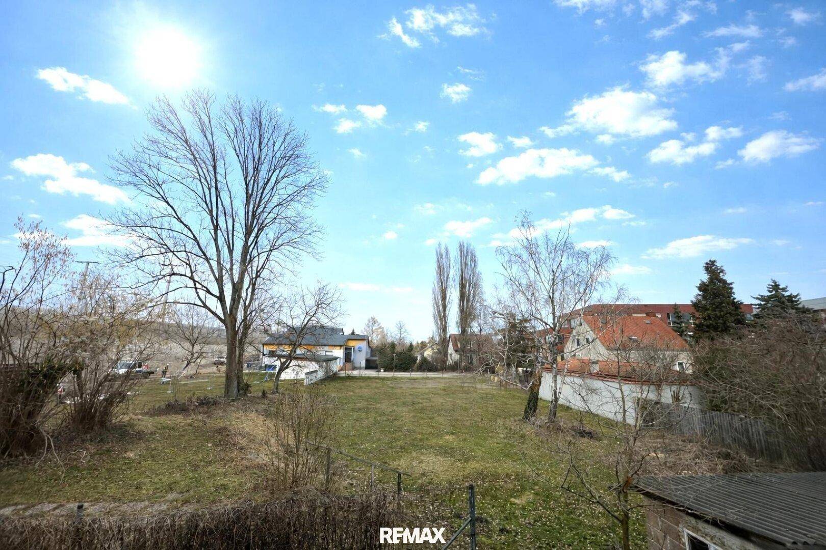 Weitläufiger Blick auf eine grüne Wiese mit Bäumen und umliegenden Häusern unter blauem Himmel.