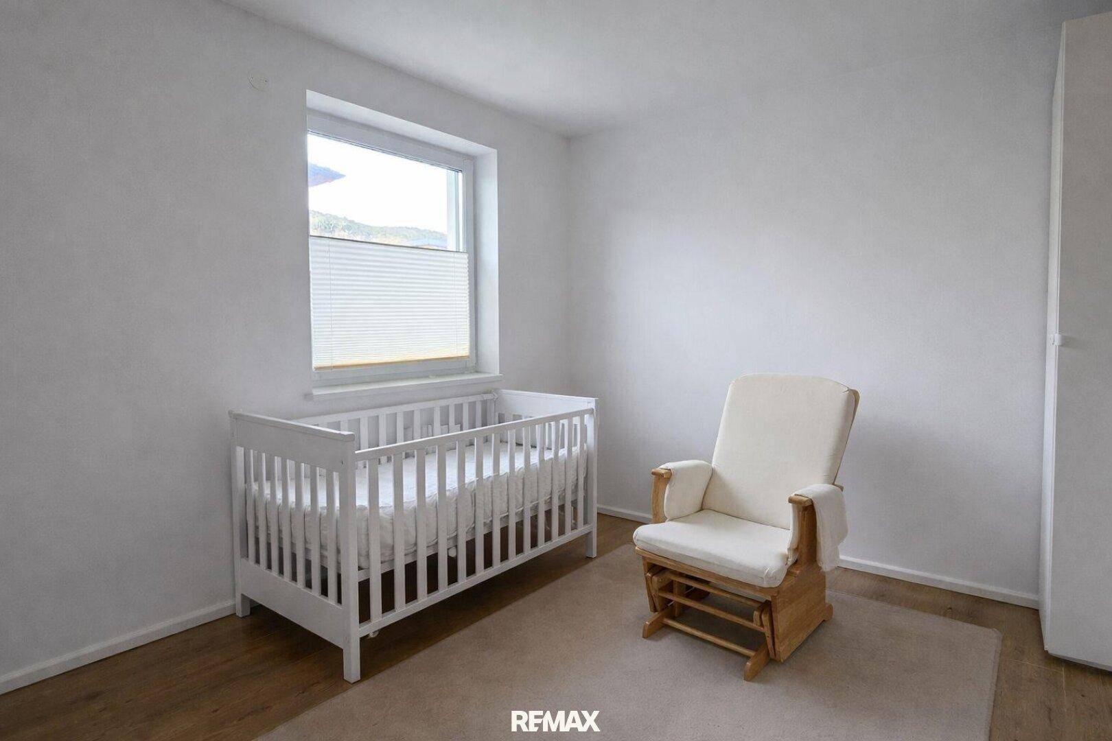 Kinderzimmer mit weißem Babybett, Schaukelstuhl und Fenster.