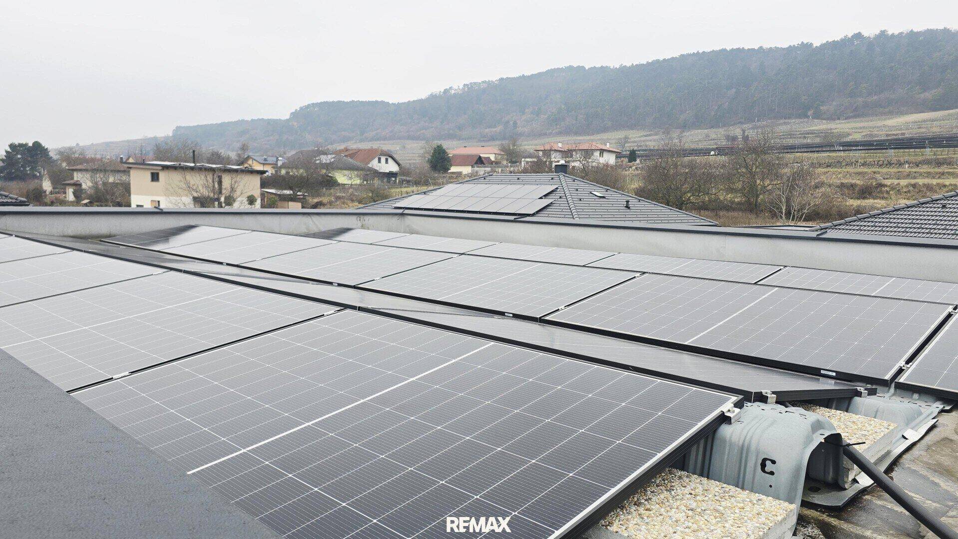 Dachfläche eines Gebäudes mit zahlreichen Photovoltaik-Modulen und hügeliger Landschaft im Hintergrund.