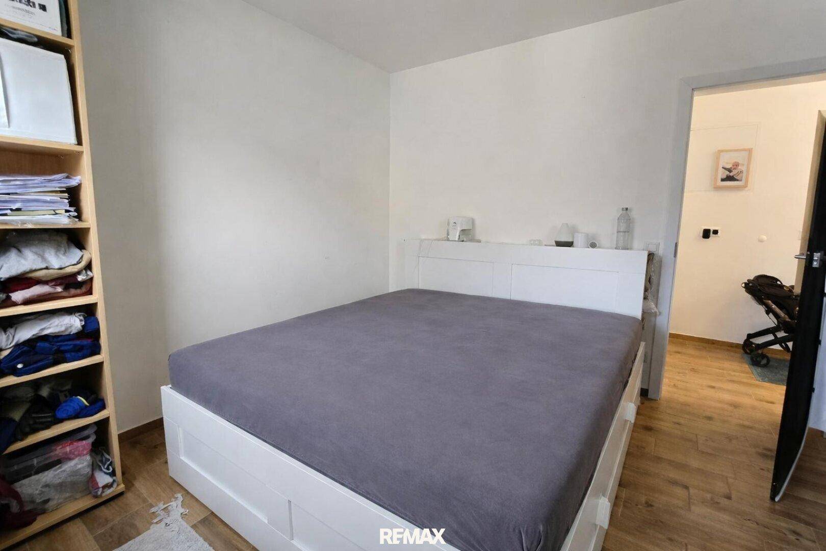 Schlafzimmer mit weißem Bettrahmen, Holzboden und offenem Zugang zu einem weiteren Raum.