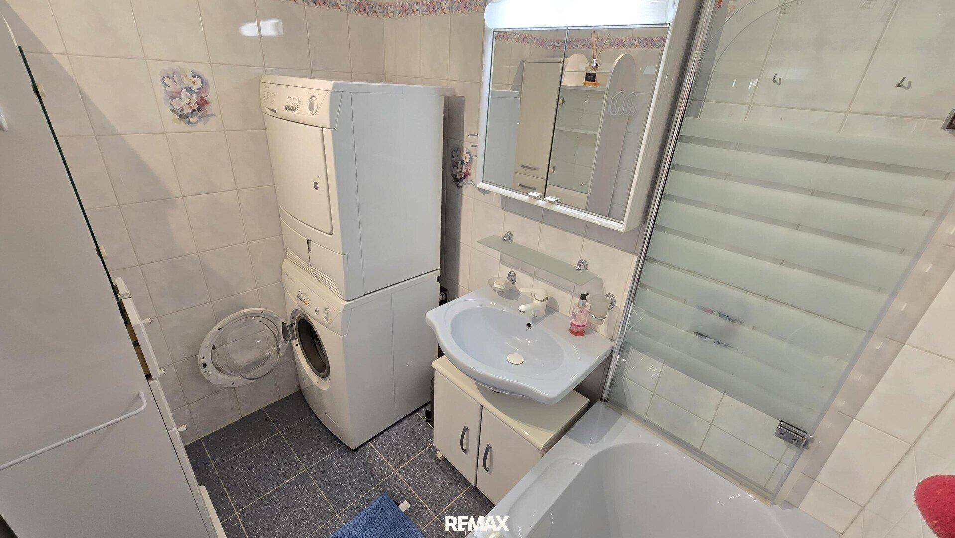 Badezimmer mit Badewanne, Waschbecken und gestapelten Haushaltsgeräten, hell gefliest.
