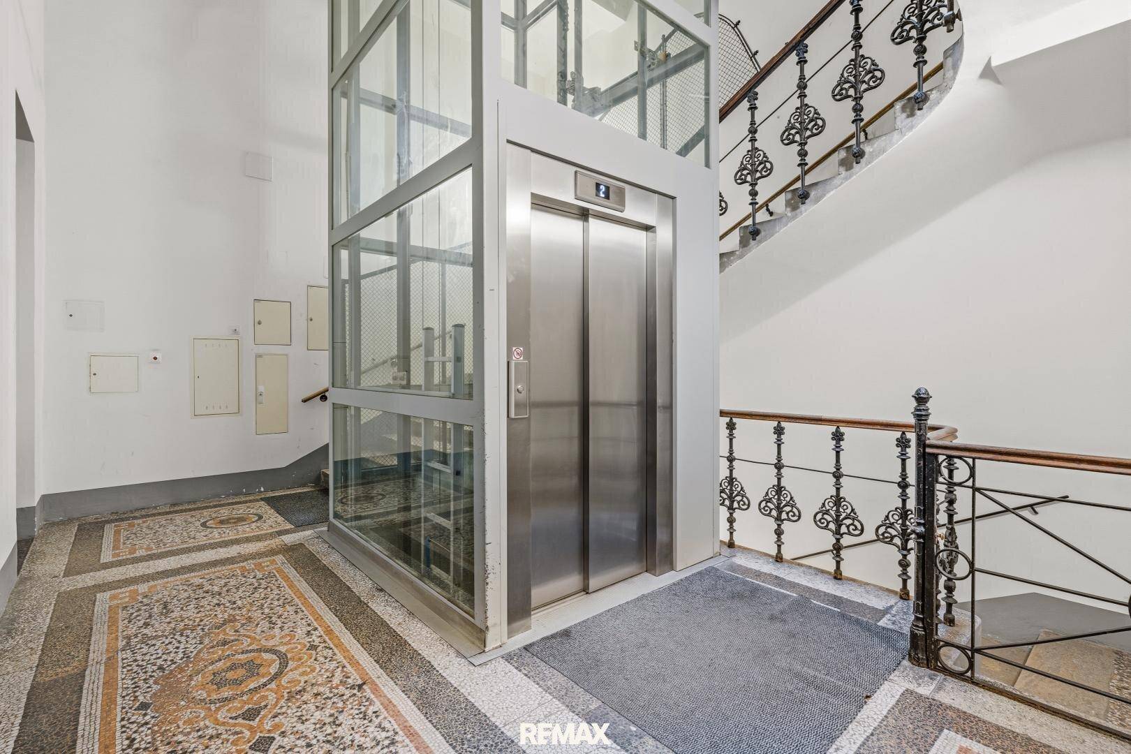 Eingangsbereich mit modernem Glasaufzug, kunstvoller Treppe und Mosaikboden.