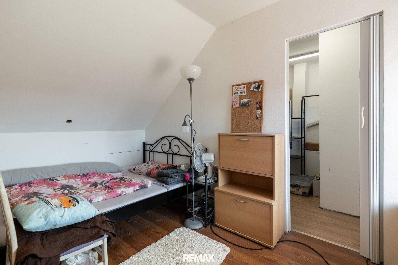 Schlafzimmer im Dachgeschoss mit Bett, Kommode und Zugang zu einem weiteren Raum.