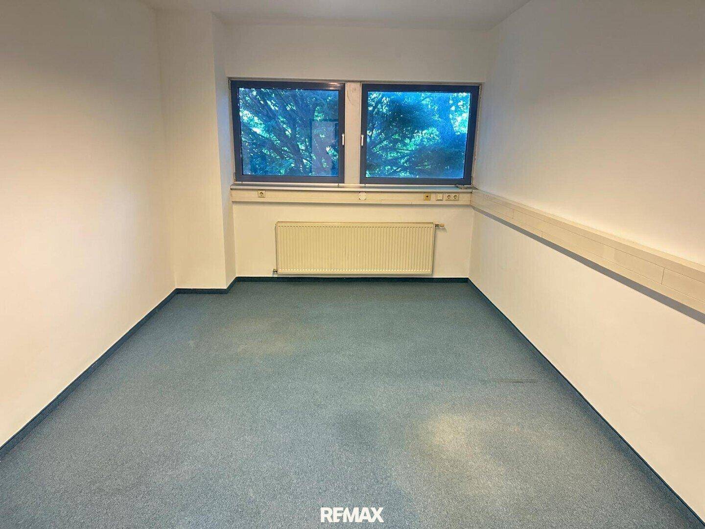 Kleineres Büro mit blauem Teppichboden, einem Heizkörper und zwei Fenstern mit Blick auf Bäume.