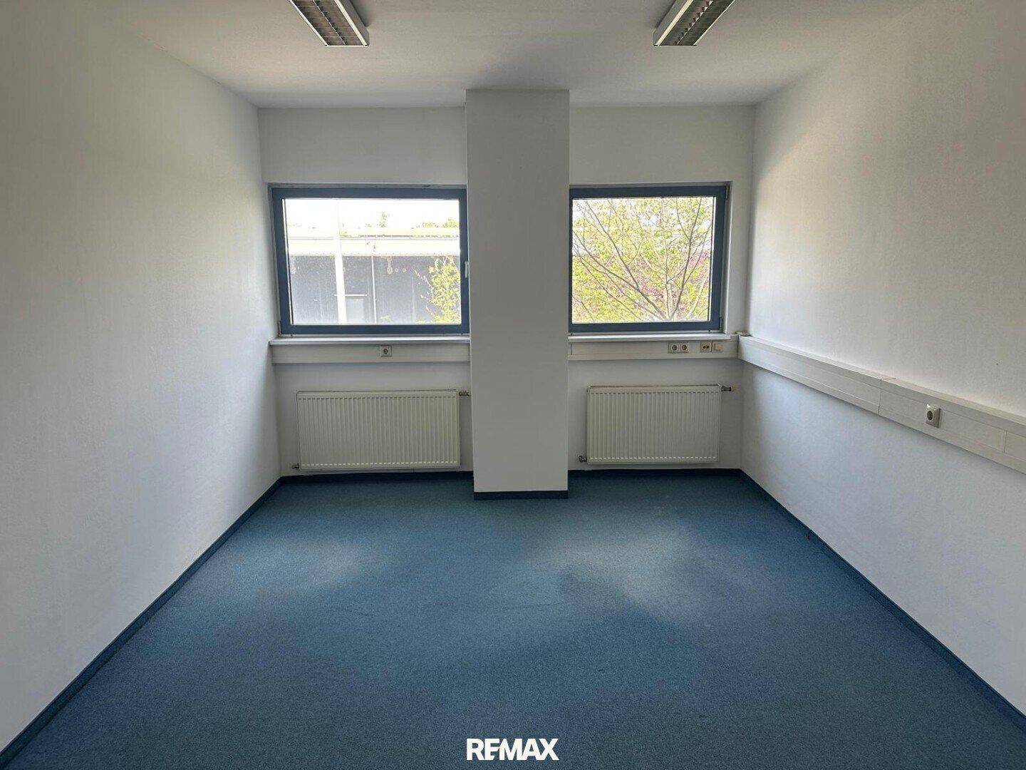 Leeres Büro mit blauem Teppichboden, einem zentralen Pfeiler und zwei Fenstern für natürliches Licht.