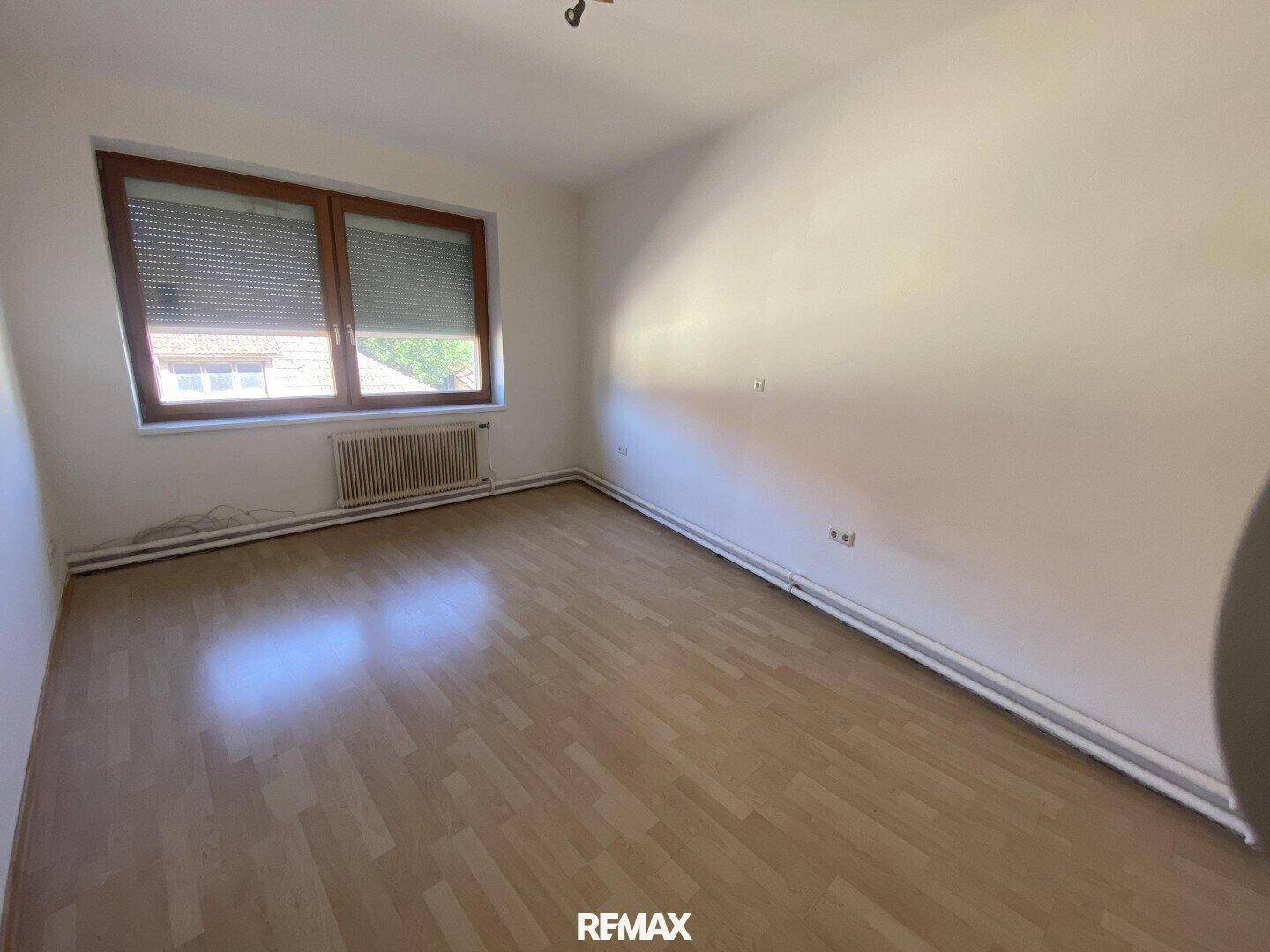 Leerer Raum mit hellem Laminatboden, weißen Wänden und einem Fenster mit Jalousien.
