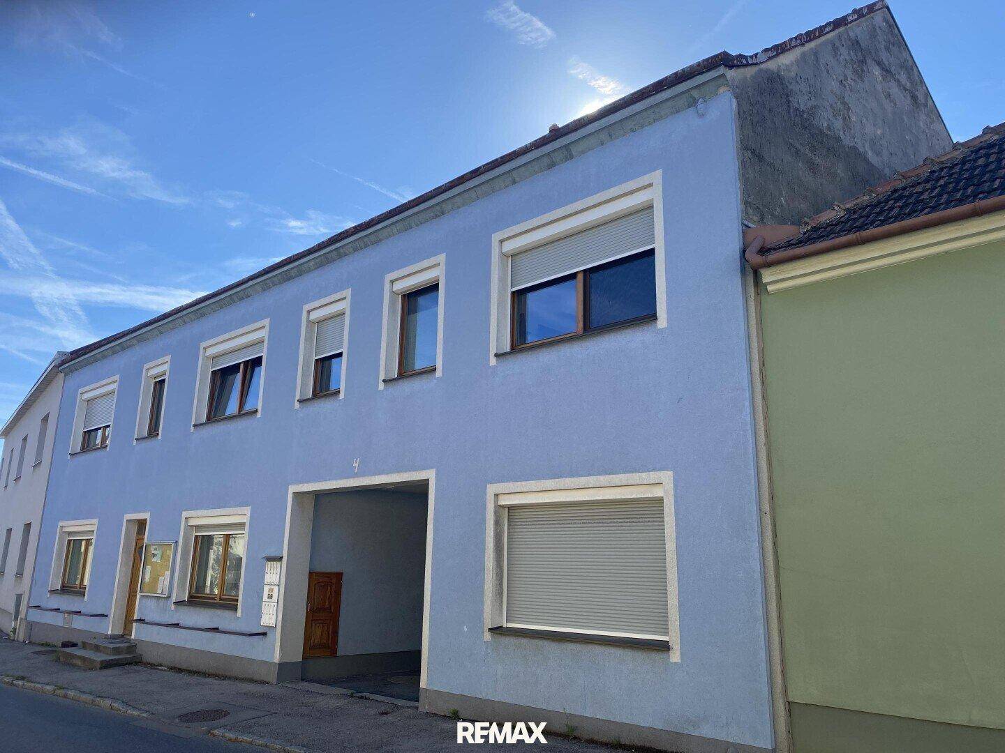 Fassade eines hellblauen Gebäudes mit mehreren Fenstern und Rollläden unter blauem Himmel.