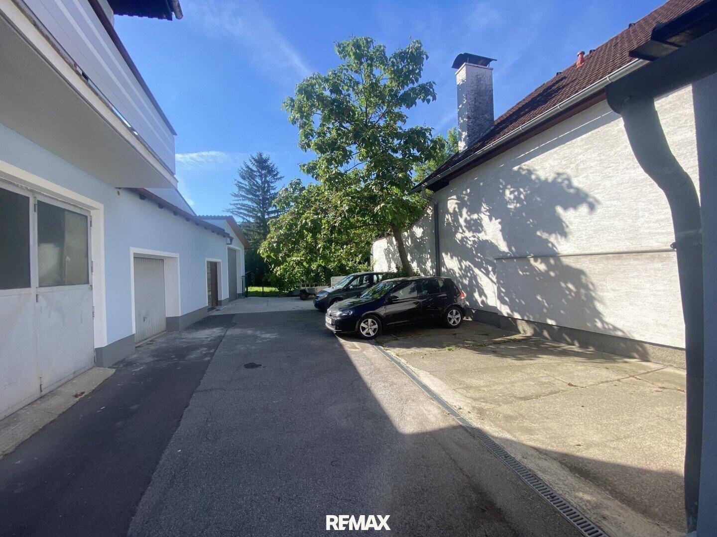 Außenbereich mit Parkplätzen zwischen Gebäuden und einem Baum im Hintergrund unter blauem Himmel.