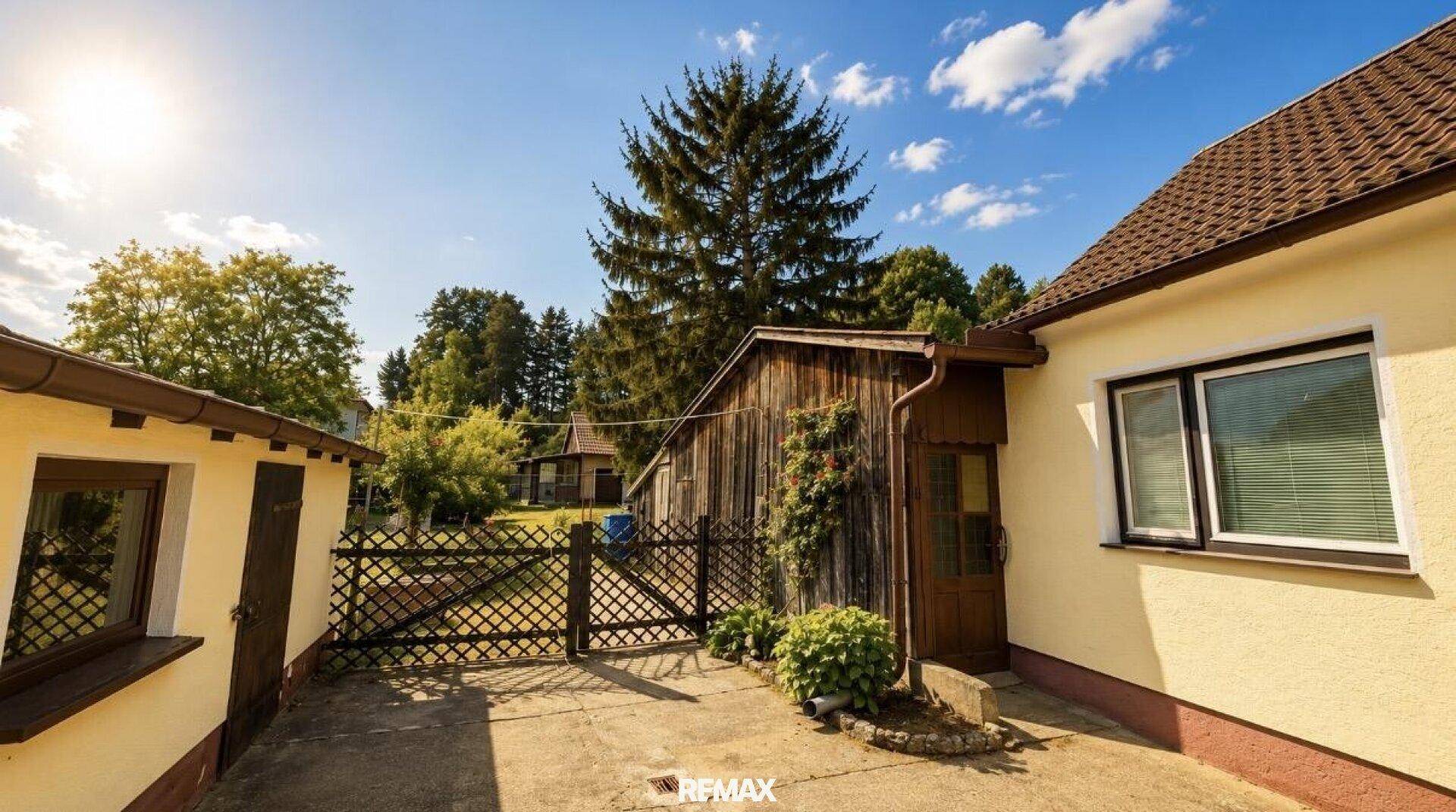 Ländliches Haus mit Holzanbau und Zaun, umgeben von Bäumen unter strahlendem Sonnenschein.