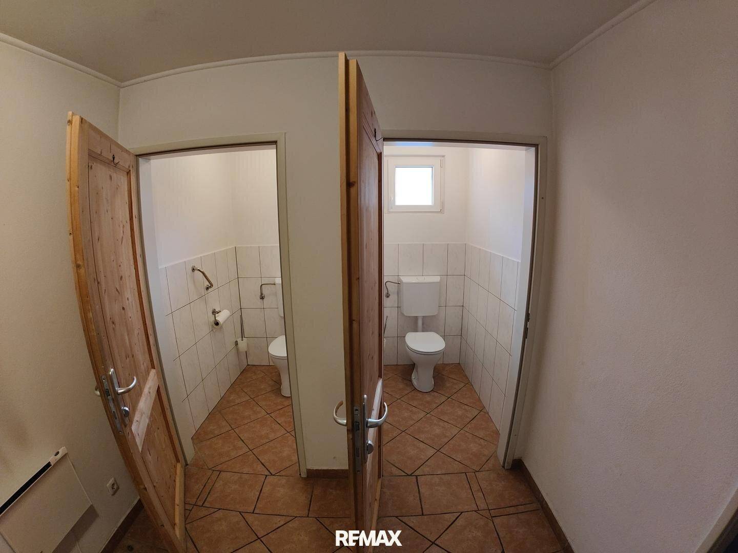 Zwei separate Toilettenräume mit Fliesenboden und weißen Wandfliesen, durch Holztüren zugänglich.