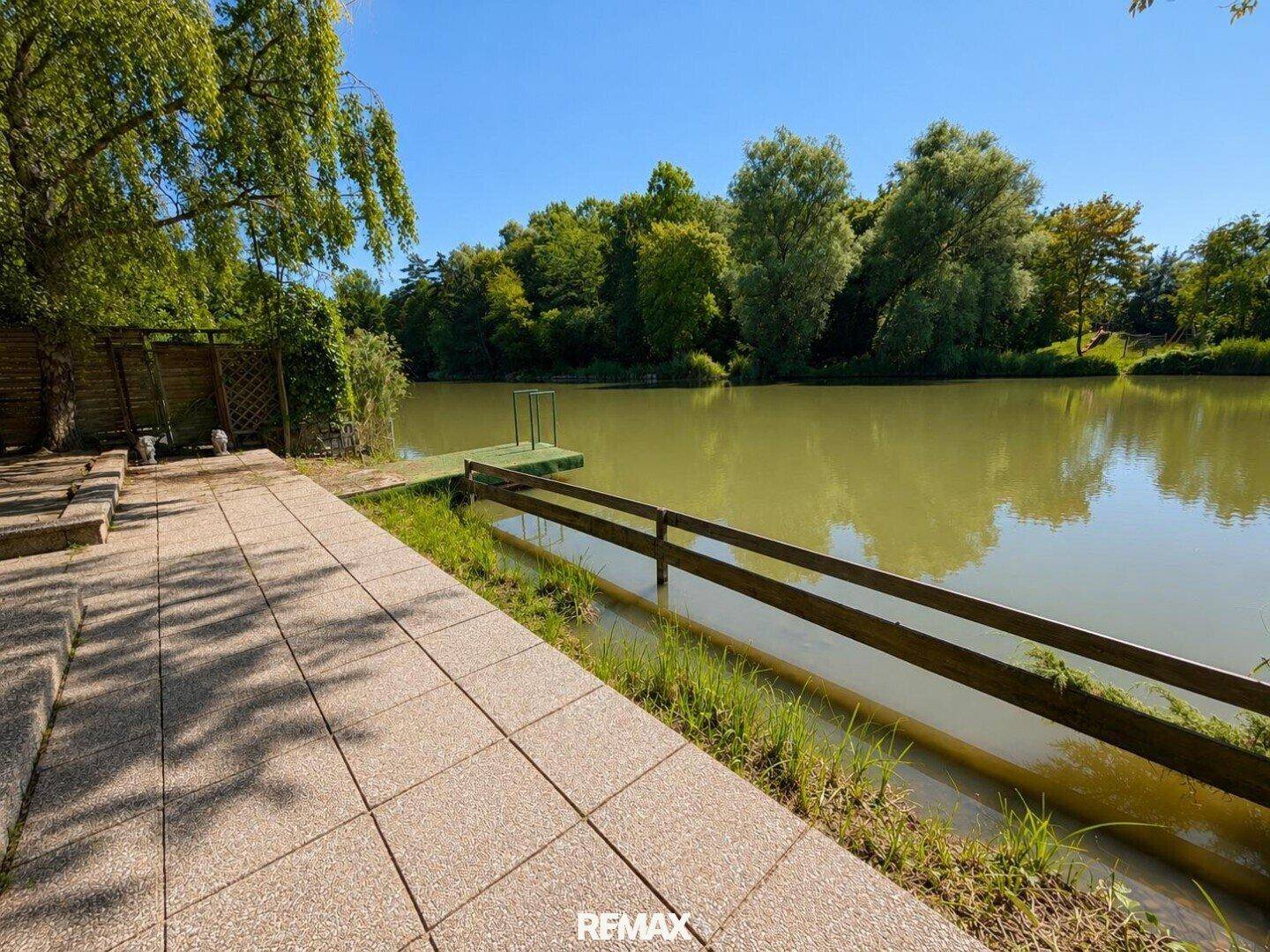 Seitlicher Blick auf den See mit einem Holzsteg und einer gepflasterten Uferpromenade.