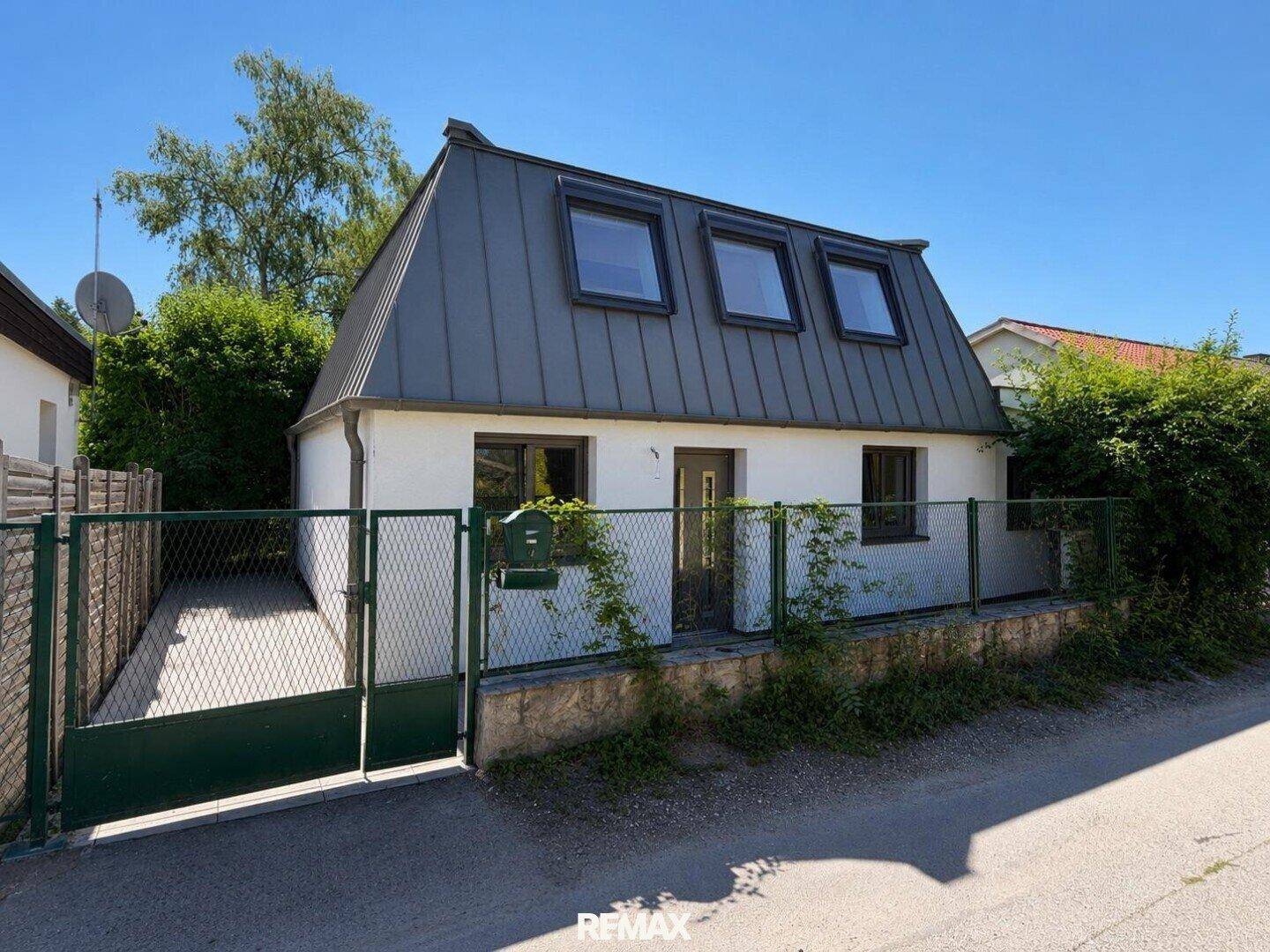 Vorderansicht eines Hauses mit Mansarddach, kleinen Fenstern und einem grünen Gartentor.