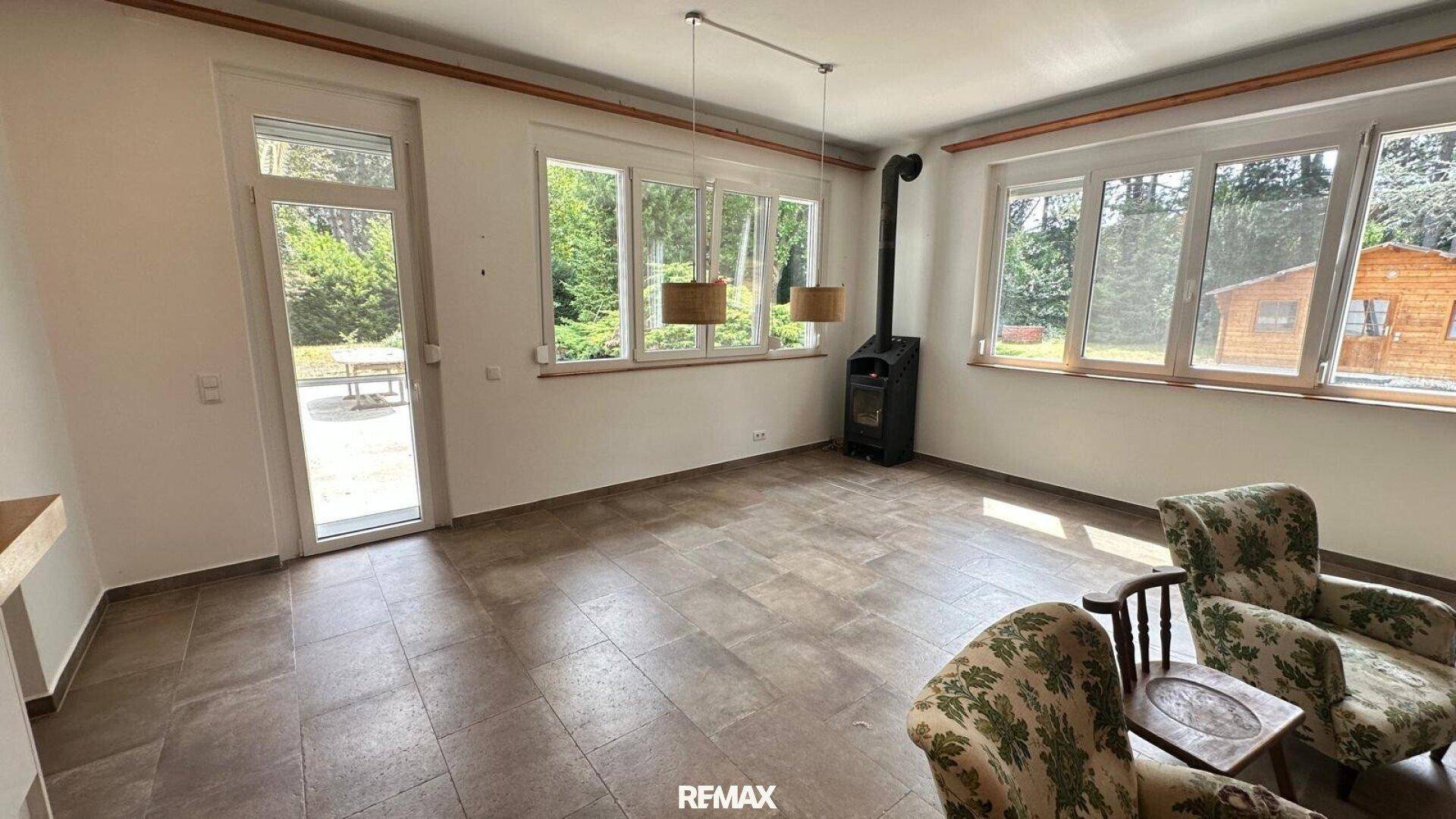 Geräumiges Wohnzimmer mit Fliesenboden, Kaminofen und großen Fenstern mit Zugang zum Garten.