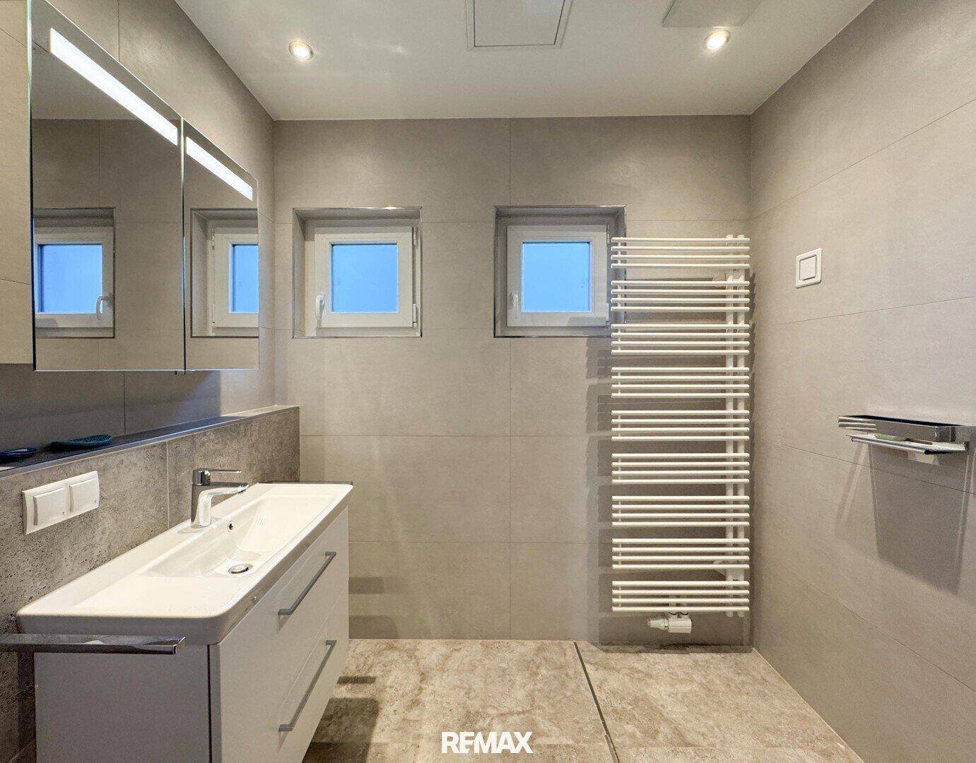 Modernes Badezimmer mit grauen Fliesen, Einzelwaschbecken, Handtuchheizkörper und zwei Fenstern.