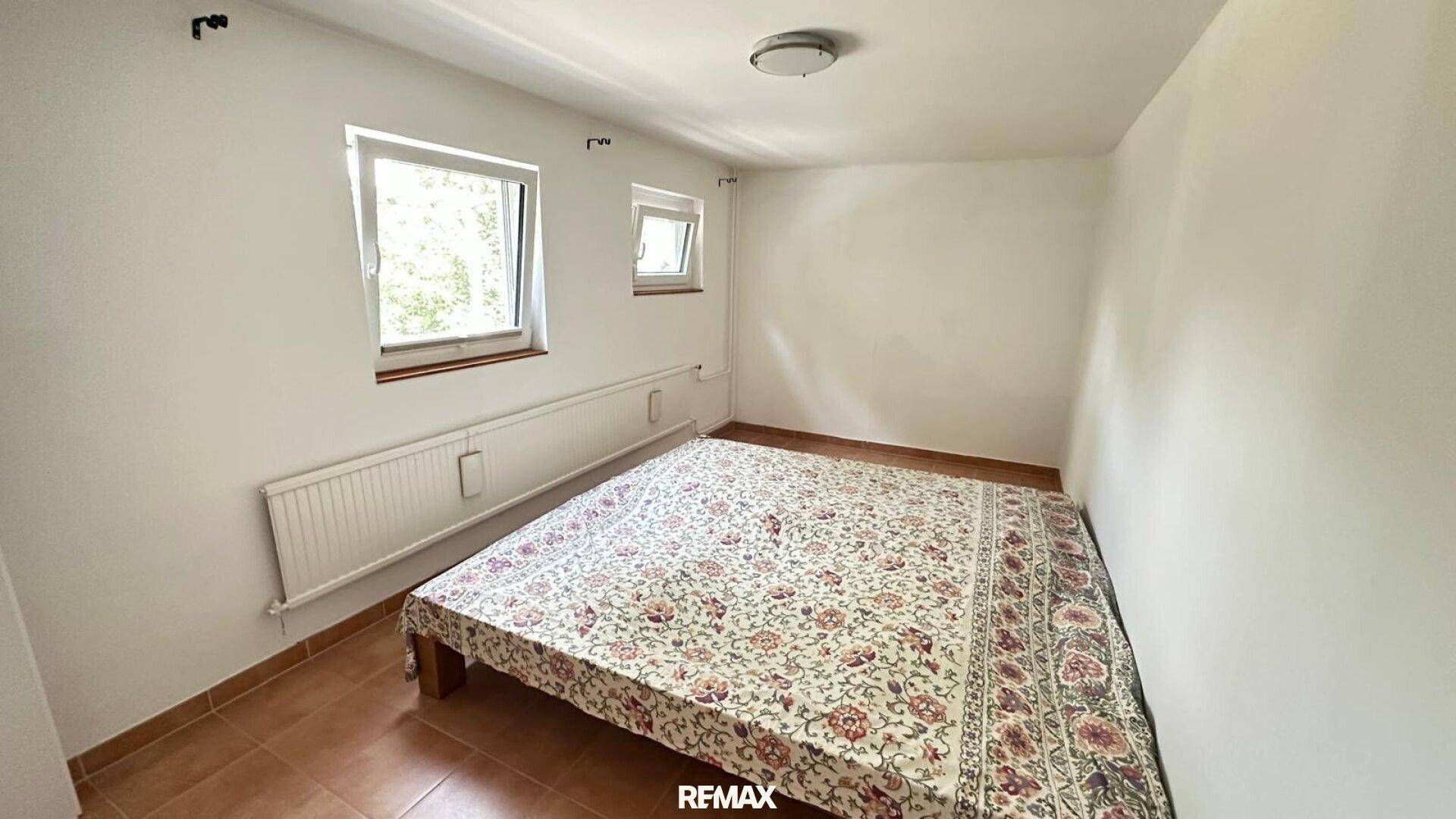 Schlafzimmer mit Fliesenboden, einem Bett und zwei Fenstern für eine helle Atmosphäre.