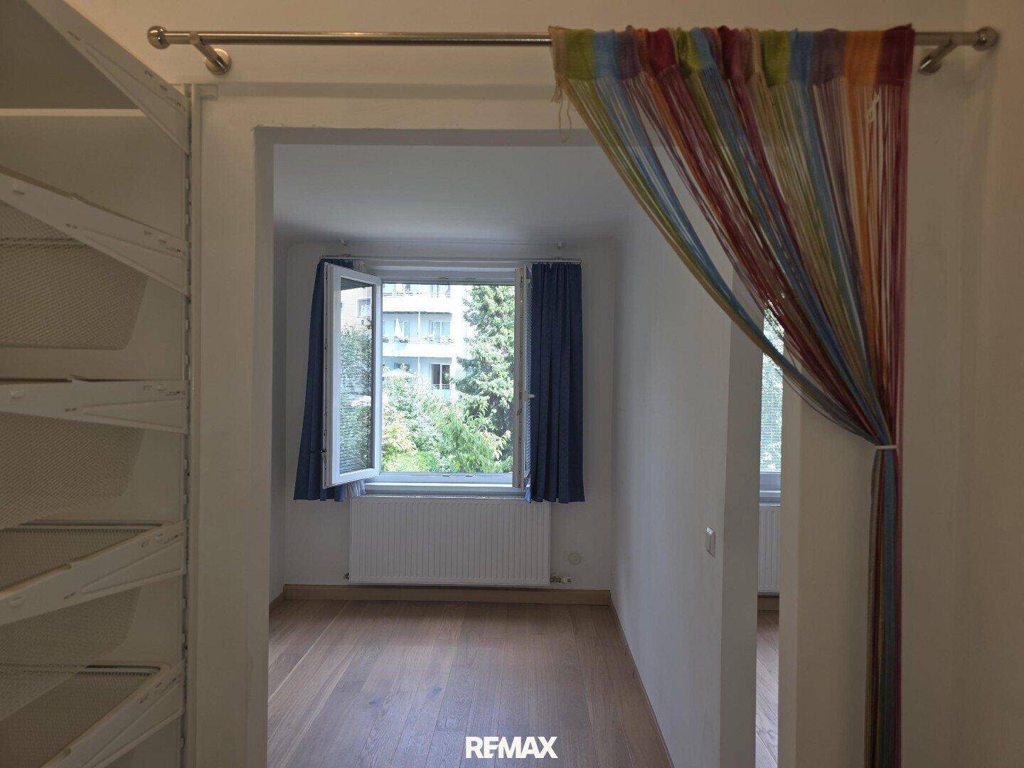 Zimmer mit Parkettboden und Fenster zum grünen Außenbereich, durch einen bunten Vorhang abgetrennt.