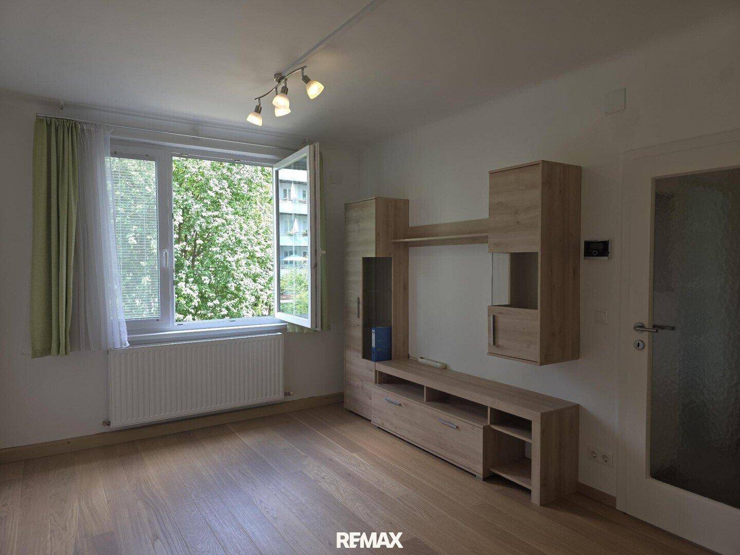 Helles Wohnzimmer mit Parkettboden, Fenster zum grünen Innenhof und einfacher Wohnwand aus Holz.