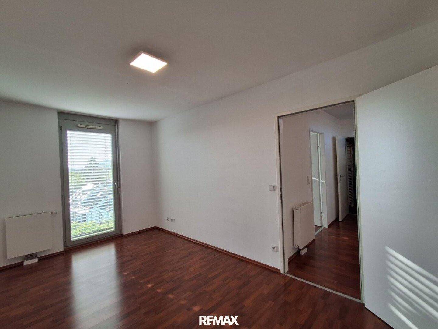 Helles Schlafzimmer mit dunklem Laminatboden und einer großen Fenstertür, die viel Licht spendet.
