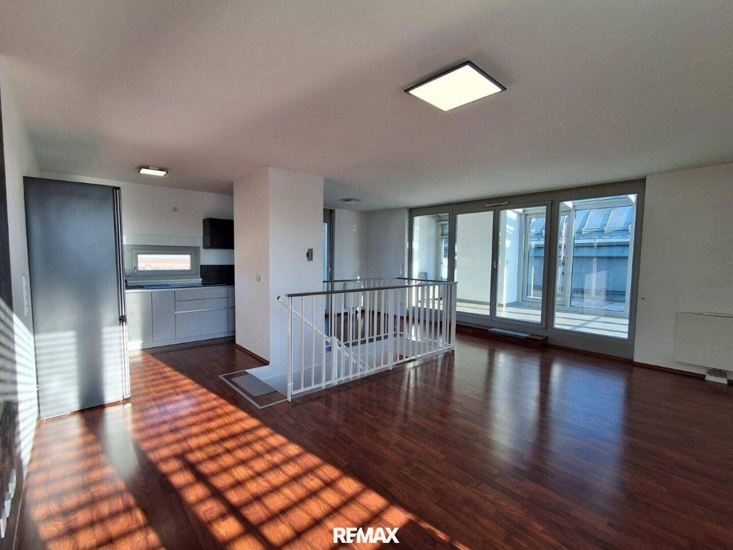 Geräumiges Wohnzimmer mit dunklem Laminatboden und großen Fenstern, die viel Tageslicht hereinlassen.