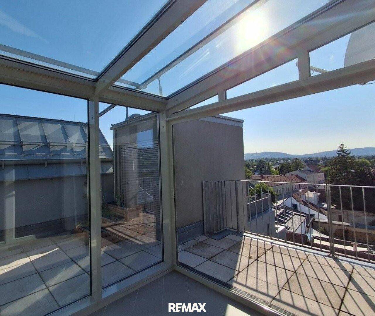 Sonniger Wintergarten mit Glasdach und Zugang zu einer gefliesten Terrasse mit weitem Ausblick.