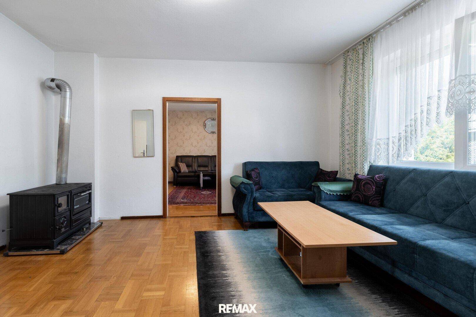 Wohnzimmer mit Holzofen, blauen Sofas und Holzboden, verbunden mit einem weiteren Raum.
