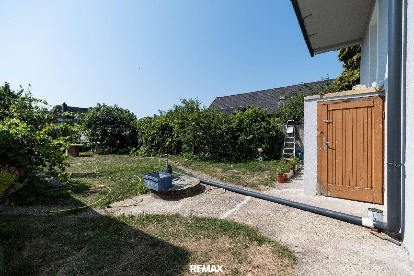 Gartenbereich mit Wasserpumpe, Schuppen und viel Grün unter blauem Himmel.