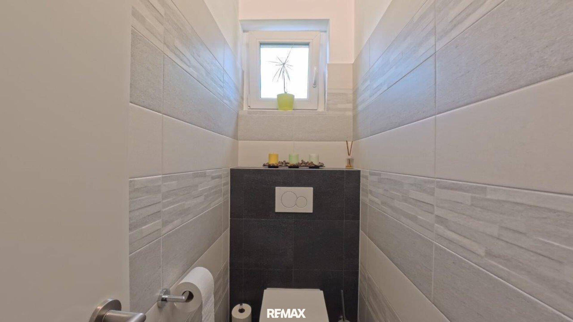 Separates WC mit Fenster, Wand-WC und modernen Fliesen in Grau und Weiß.