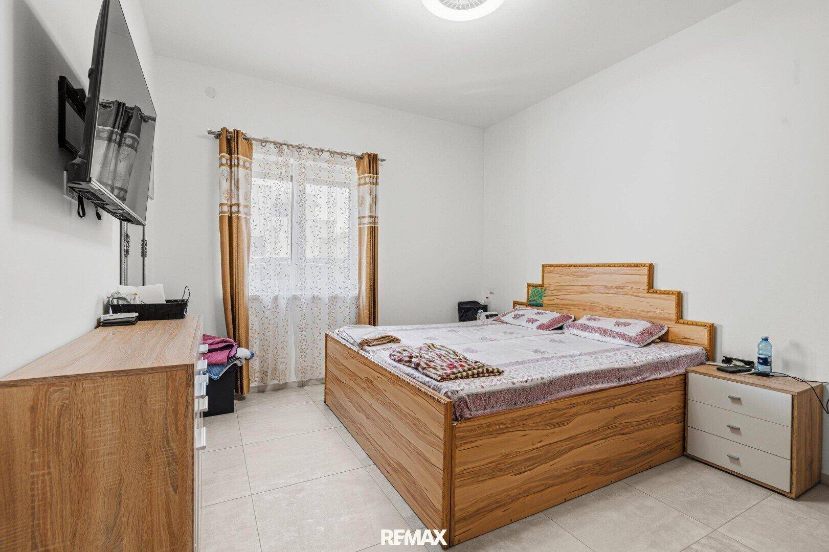 Schlafzimmer mit Doppelbett aus Holz, Fliesenboden und Fenster mit Vorhängen.