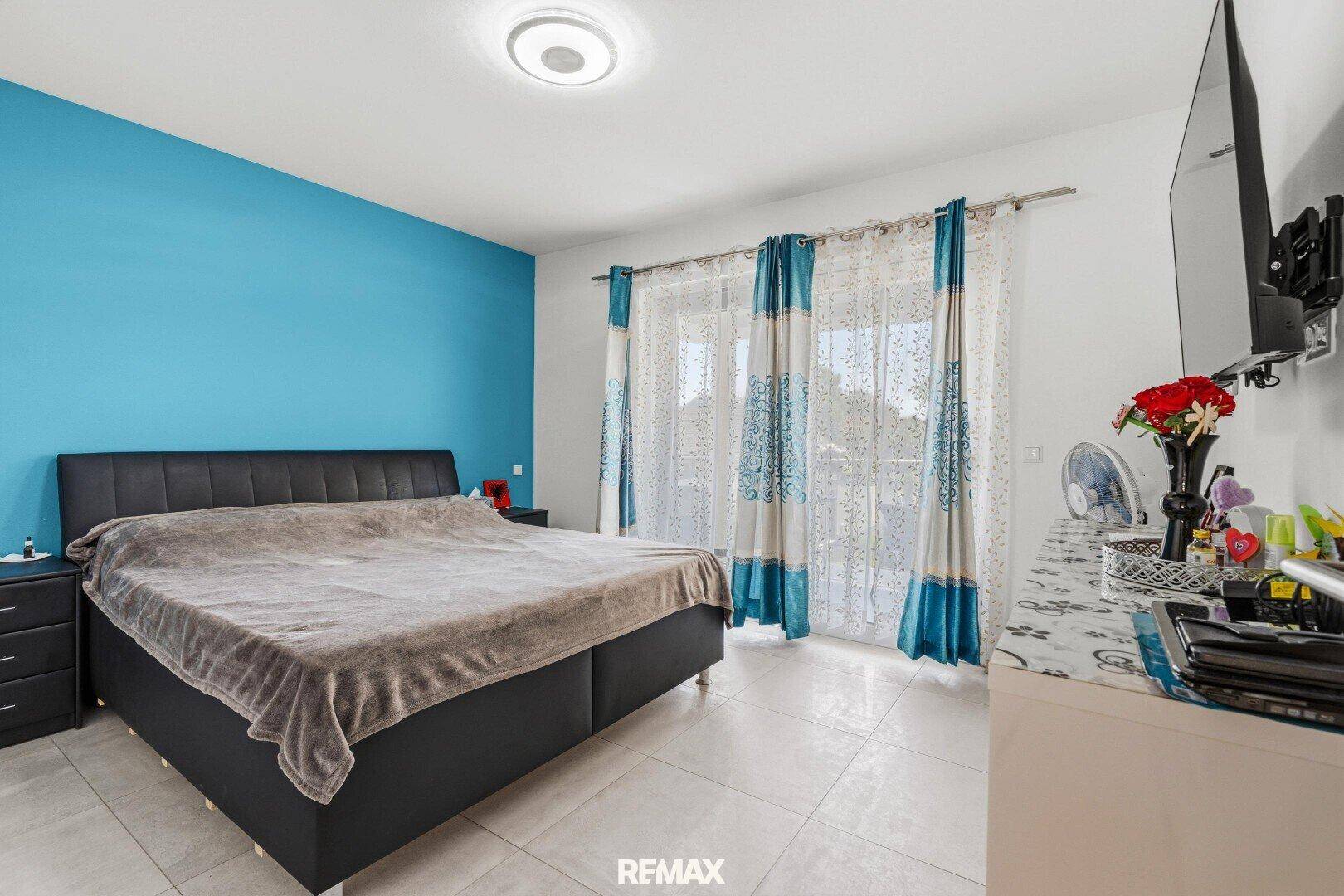Helles Schlafzimmer mit blau akzentuierter Wand, Doppelbett und Zugang zum Balkon.