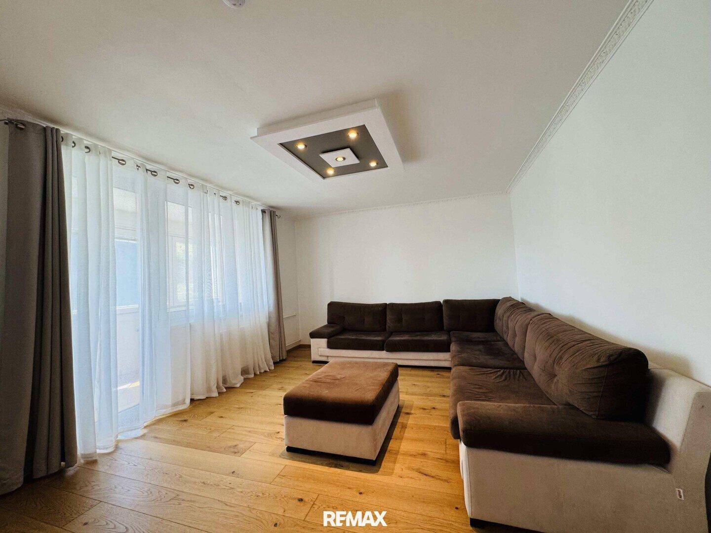 Geräumiges Wohnzimmer mit brauner Eckcouch, Holzboden und großem Fenster mit Zugang nach draußen.