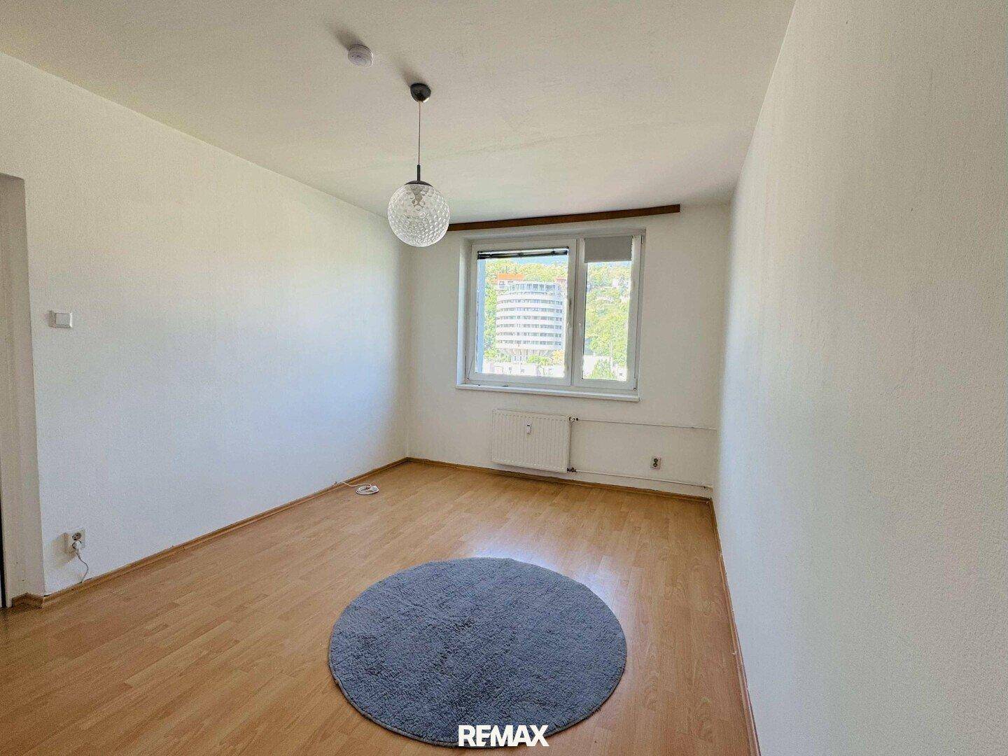 Helles, leeres Zimmer mit Holzboden, weißen Wänden und einem Fenster mit Blick auf die Stadt.