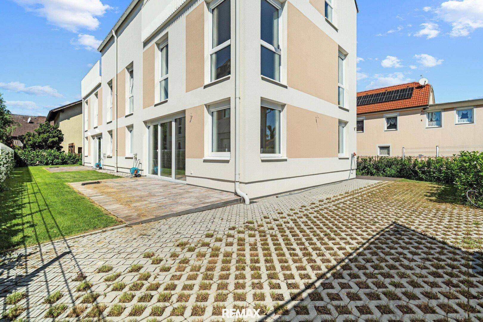 Gartenbereich mit Rasen, gepflasterter Terrasse und Parkflächen, umgeben von Hecken.