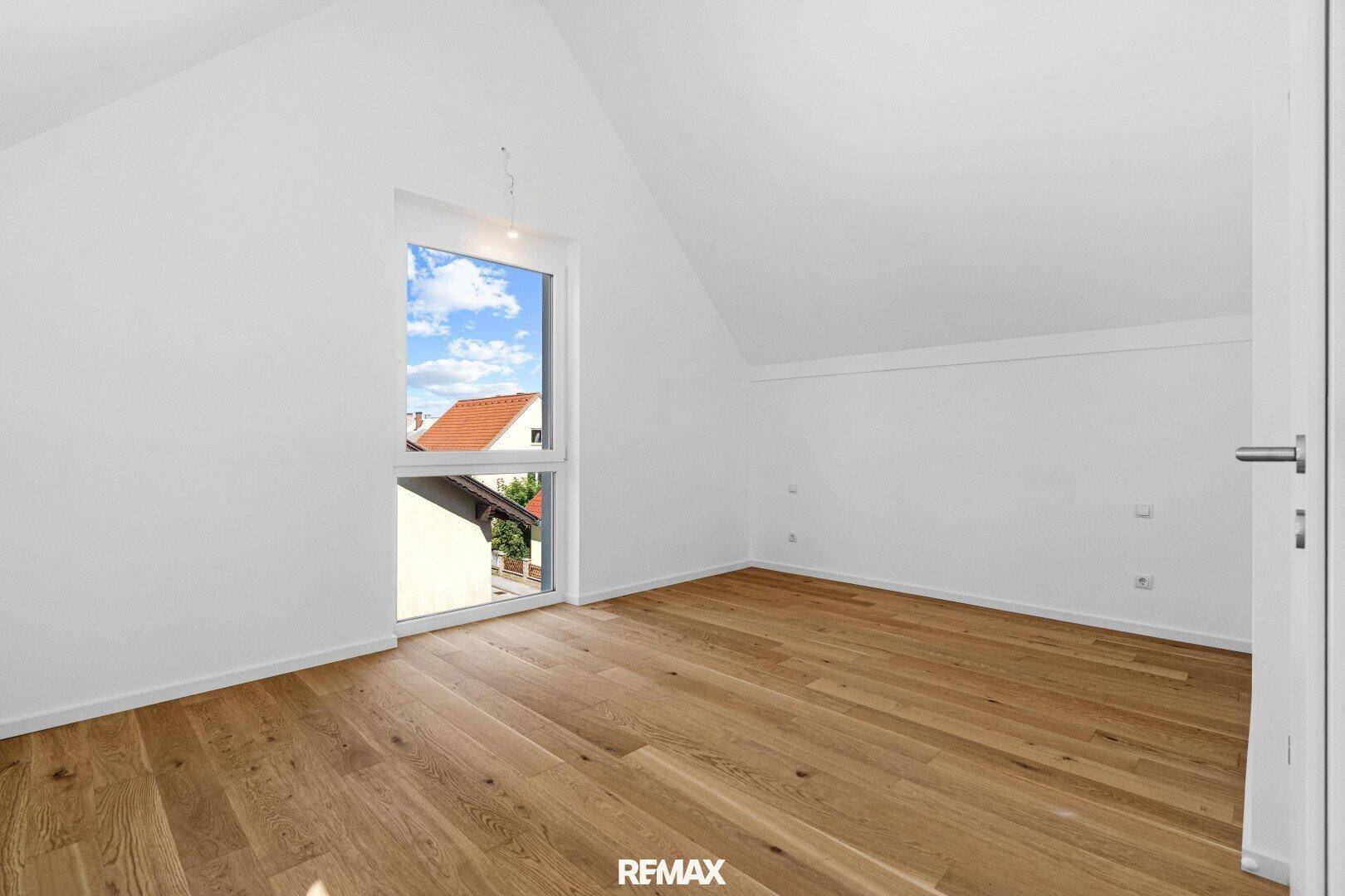 Dachzimmer mit schrägen Wänden, Holzboden und einem Fenster mit Blick auf die Umgebung.
