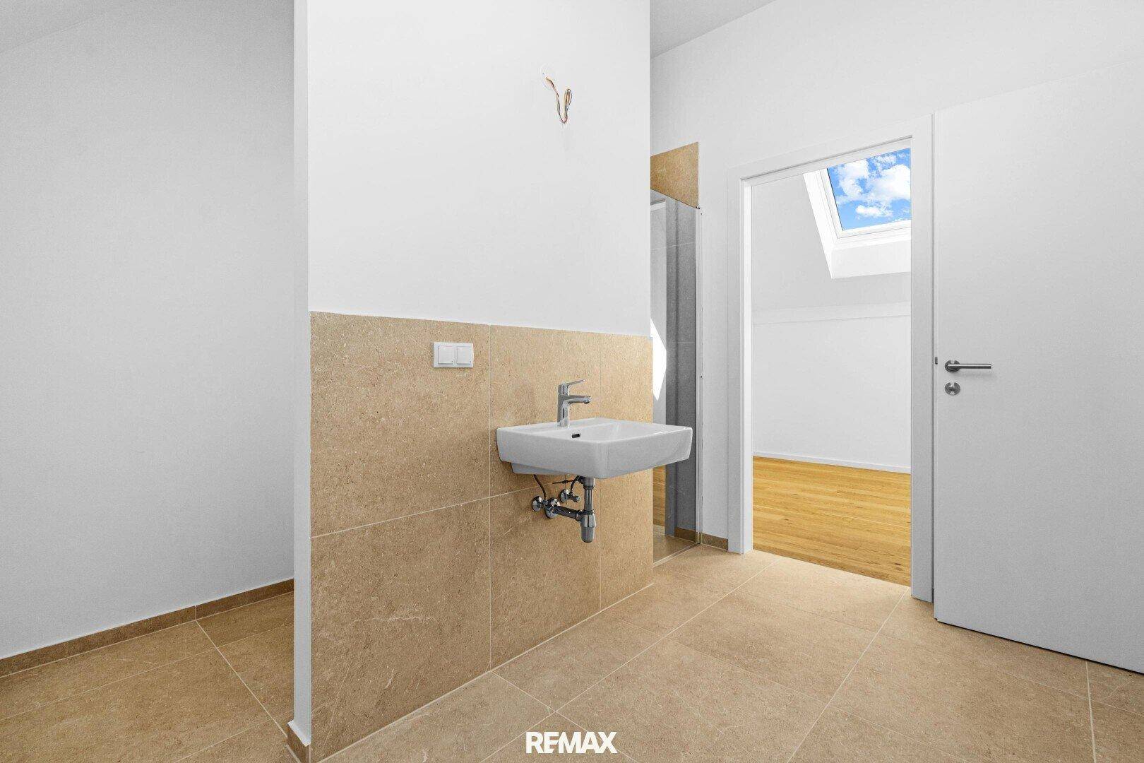Badezimmerbereich mit Waschbecken, beigefarbenen Fliesen und Blick in einen Raum mit Dachfenster.