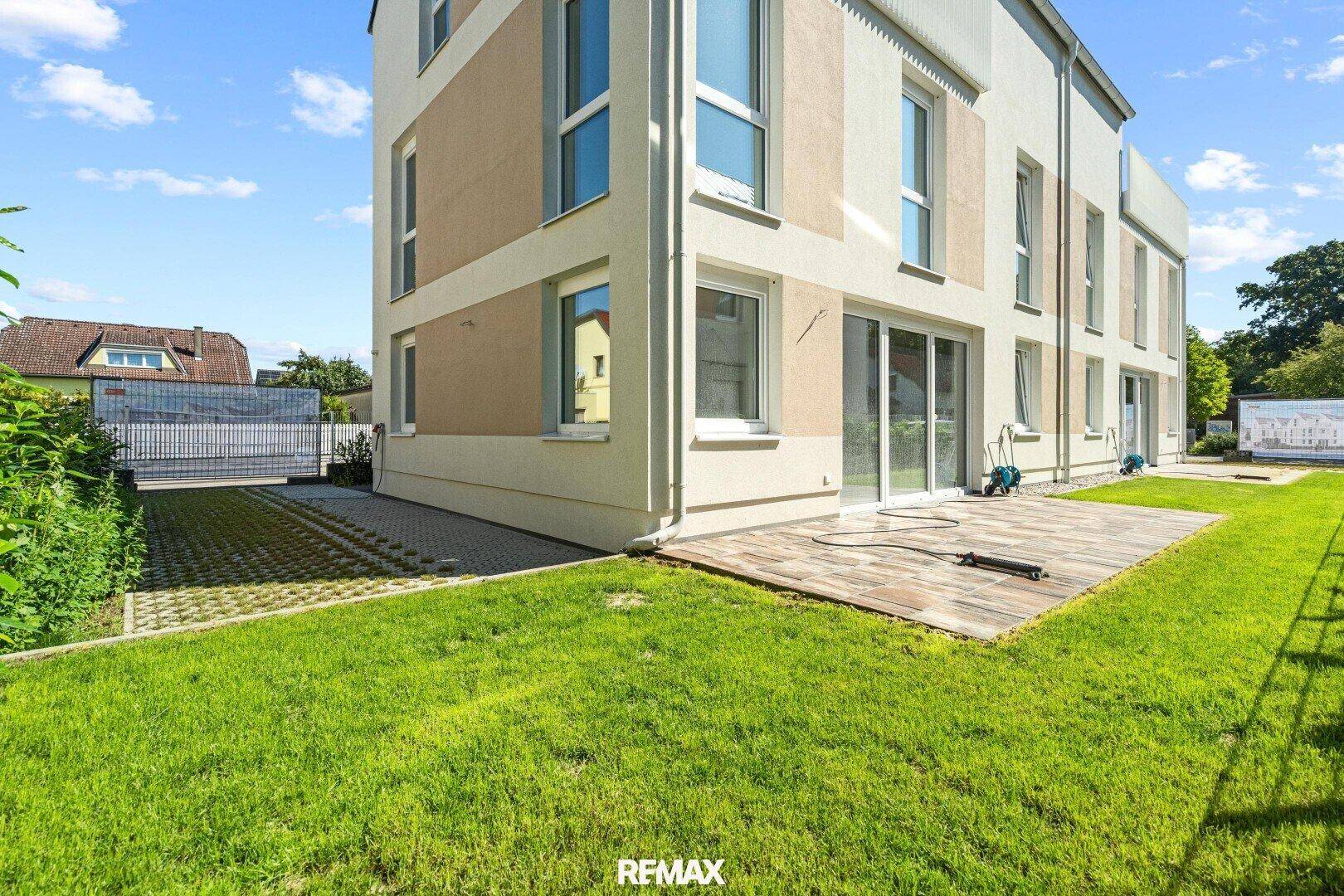 Seitlicher Gartenbereich mit Rasenfläche, Holzterrasse und gepflasterter Zufahrt zum Haus.
