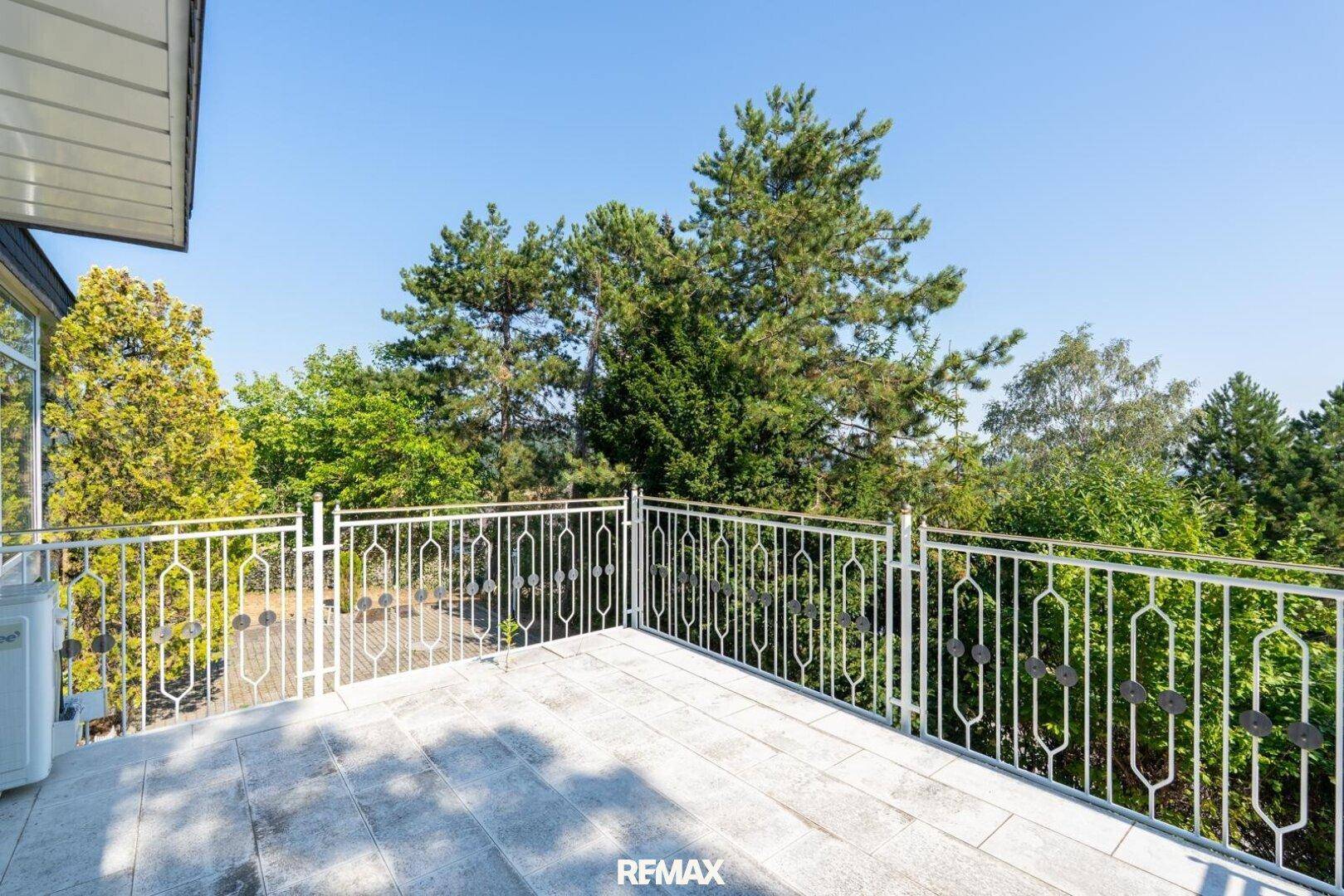 Großer Balkon mit Fliesenboden und schmiedeeisernem Geländer, umgeben von Bäumen.