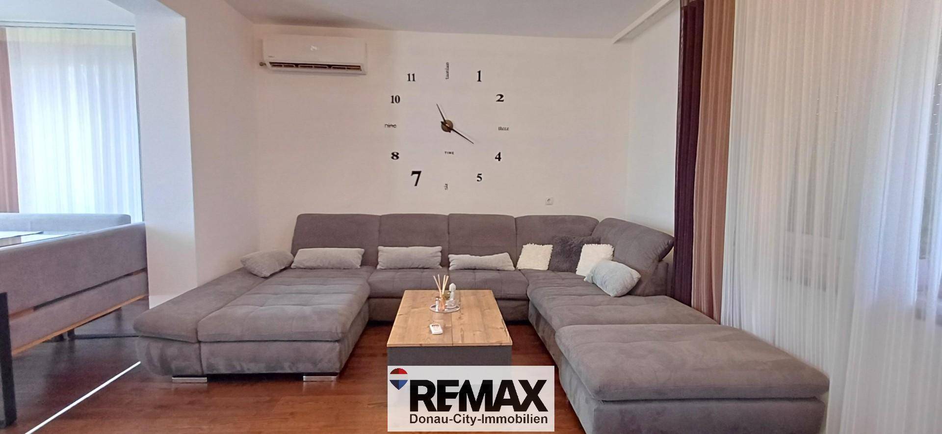 Geräumiges Wohnzimmer mit großem Ecksofa, Holzboden und moderner Wanduhr.