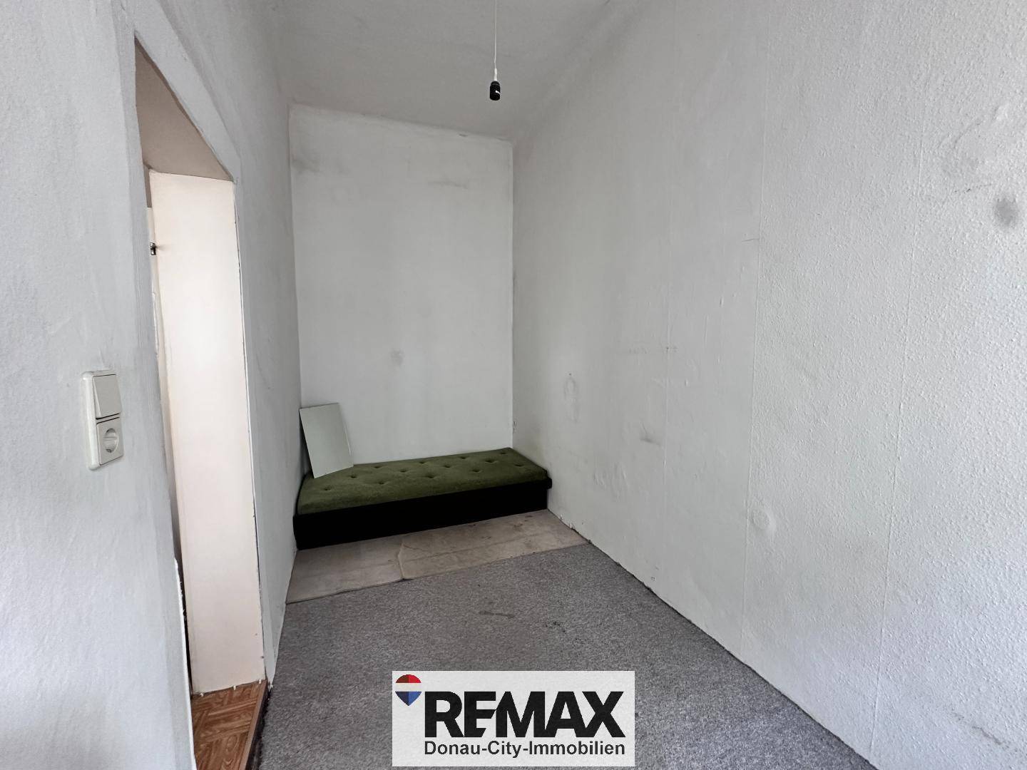 Kleines, einfaches Zimmer mit grauem Teppichboden und einer grünen Matratze.