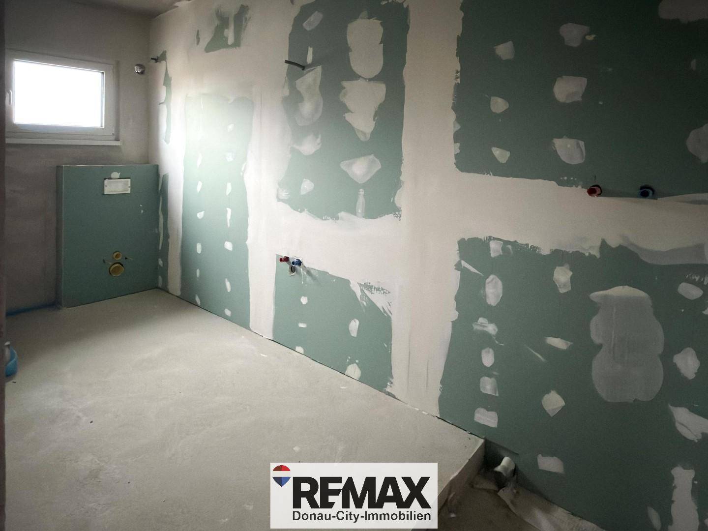 Unfertiges Badezimmer im Rohbau mit Fenster und vorbereiteten Installationen.